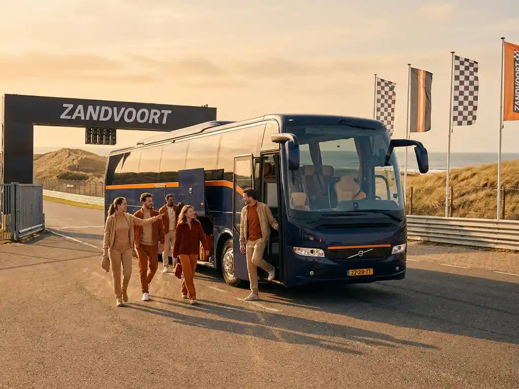 Luxe touringcar bij de ingang van Circuit Zandvoort, passagiers stappen uit op zonnig asfalt met duinen en zee op de achtergrond.