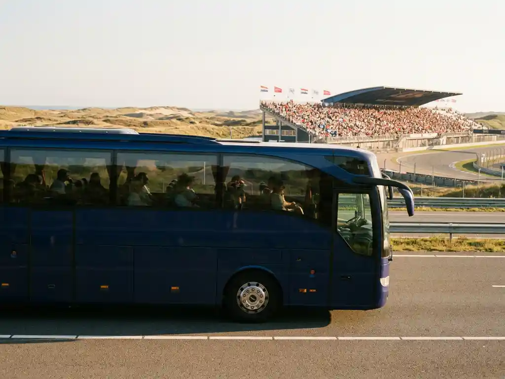 Luxe touringcar op lege Nederlandse snelweg bij Zandvoort, met kustduinen en gevulde racetribunes op de achtergrond.