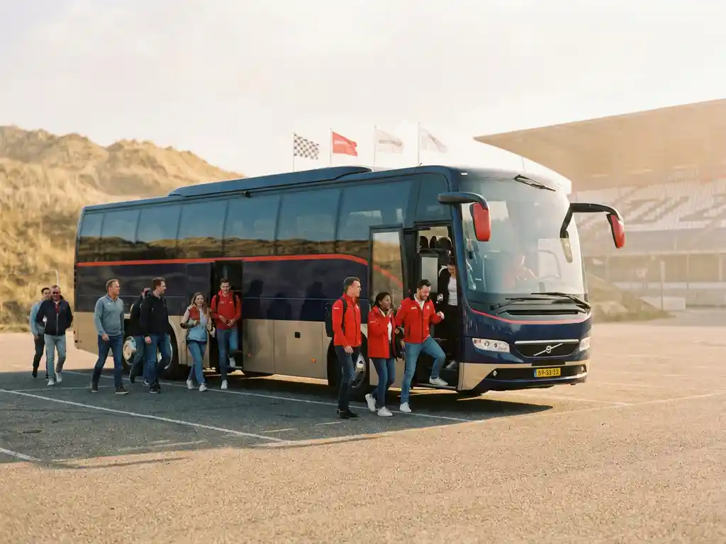 Luxe touringcar bij Circuit Zandvoort met passagiers die uitstappen, racevlaggen en duinen op de achtergrond in warm kustlicht.